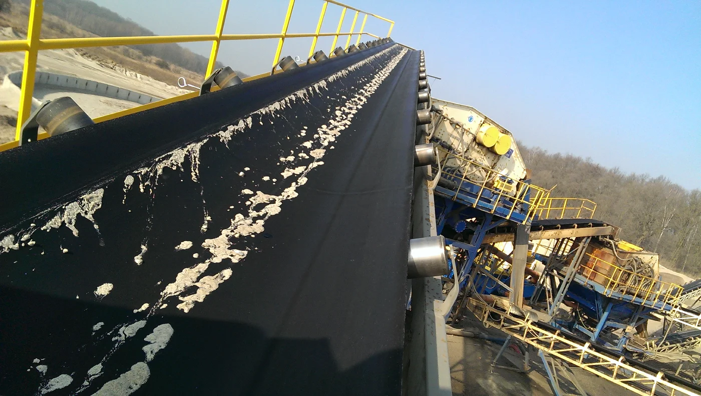 Large panoramic photo showing a long, black conveyor belt stretching diagonally across the center of the frame, viewed from above. The conveyor is slightly soiled with light, irregular sand stains. To the left of the belt, along its edge, there is a yellow metal safety barrier with a handrail, running upwards and to the right. To the right of the belt, metal support rollers are visible. In the background, to the right of the conveyor, there is a large, complex industrial machinery structure in yellow and blue, used for crushing materials. In the distance to the right of the conveyor, trees and a fragment of a forest are visible, while to the left of the horizon, an open mining area with visible sand and rubble heaps can be seen. The entire scene is illuminated by bright daylight, and the sky is clear and blue.