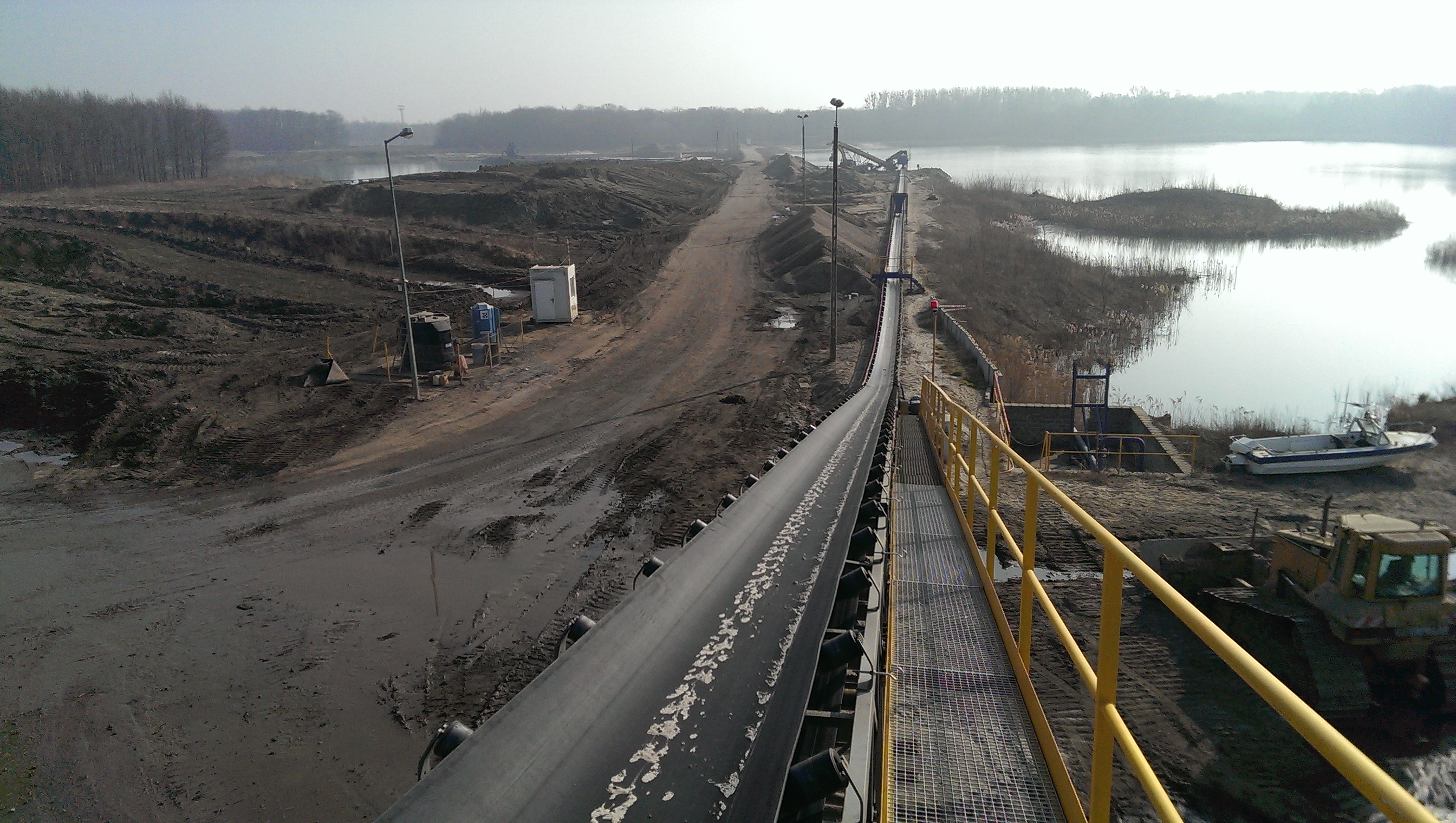 Large panoramic photo showing a long, black conveyor belt stretching diagonally across the center of the frame, viewed from above. The conveyor is slightly soiled with light, irregular sand stains. To the left of the belt, along its edge, there is a yellow metal safety barrier with a handrail, running upwards and to the right. To the right of the belt, metal support rollers are visible. In the background, to the right of the conveyor, there is a large, complex industrial machinery structure in yellow and blue, used for crushing materials. In the distance to the right of the conveyor, trees and a fragment of a forest are visible, while to the left of the horizon, an open mining area with visible sand and rubble heaps can be seen. The entire scene is illuminated by bright daylight, and the sky is clear and blue.